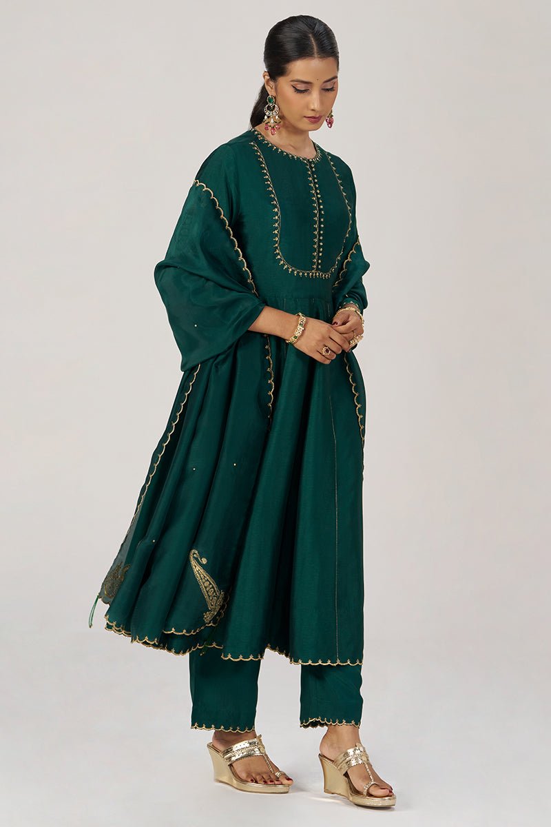 A woman modeling a dark green traditional outfit with gold embroidered details, paired with matching pants and gold wedge sandals, against a light gray studio background. | Emerald Green Raw Silk Festive Kurta Set - Ganga Fashions