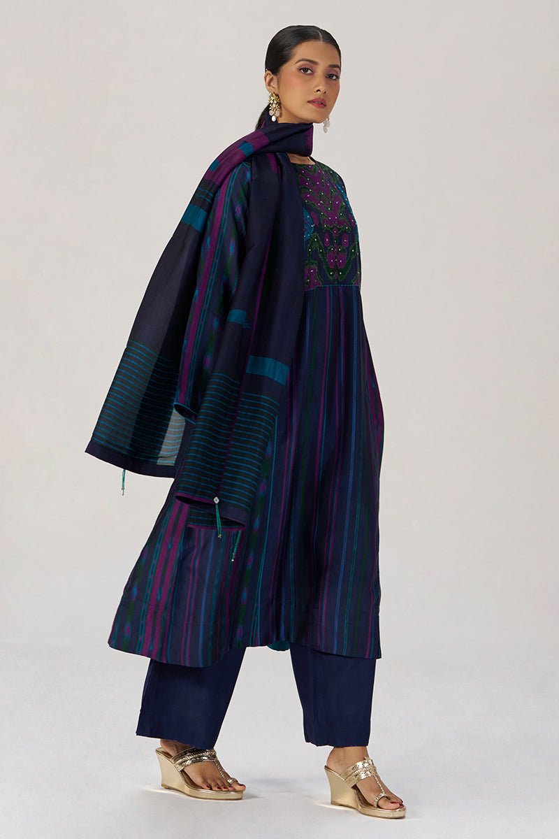 A model wearing a dark navy blue traditional outfit with vertical stripes in purple, teal, and green, featuring a matching embroidered tunic, wide-leg pants, and a draped scarf, set against a plain light gray background. | Medieval Blue Silk Festive Kurta Set - Ganga Fashions