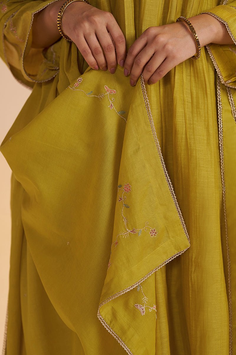 A close-up of a person wearing a mustard yellow traditional garment with delicate pink floral embroidery and a thin metallic embroidered border. The person's hands and arms are visible, adorned with gold-toned bangles, against a solid light beige background. | Lemon Curry Festive Kurta Set - Ganga Fashions