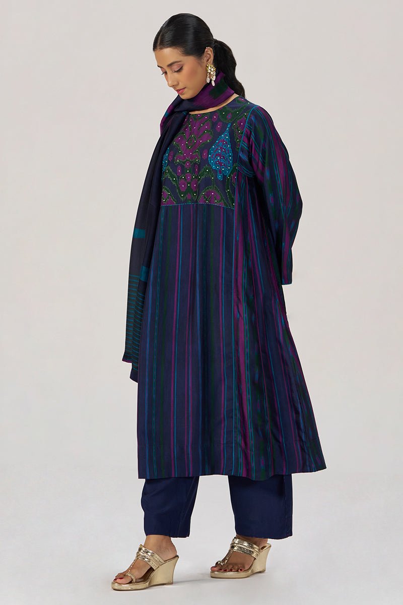 A woman models a dark navy blue embroidered kurta set with vertical stripes in purple and green, paired with matching navy pants and a navy scarf, against a light gray studio background. | Medieval Blue Silk Festive Kurta Set - Ganga Fashions