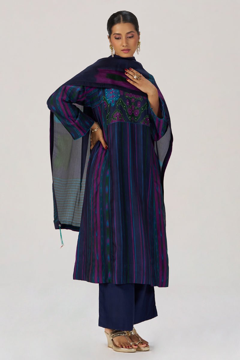 A woman models a traditional Indian outfit consisting of a navy blue kurta with purple and green vertical stripes, a matching navy blue dupatta, and navy blue pants. The kurta features a heavily embroidered bodice with floral patterns in purple, green, and gold. | Medieval Blue Silk Festive Kurta Set - Ganga Fashions