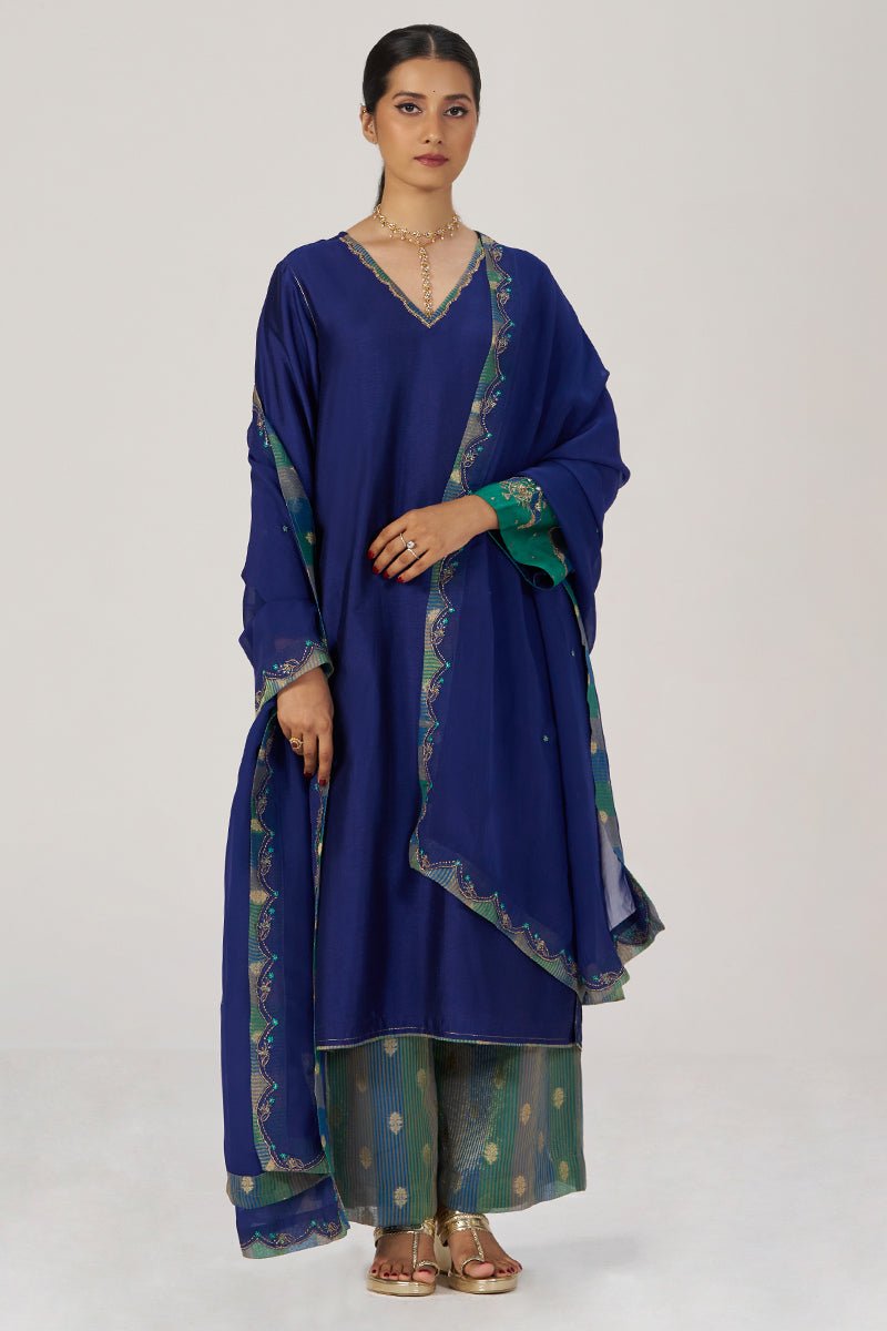A woman modeling a traditional Indian outfit consisting of a royal blue kurta with green and gold border details, matching green pants with gold motifs, and a blue dupatta draped over her shoulder, set against a plain light gray studio background. | Space Blue Festive Kurta Set - Ganga Fashions