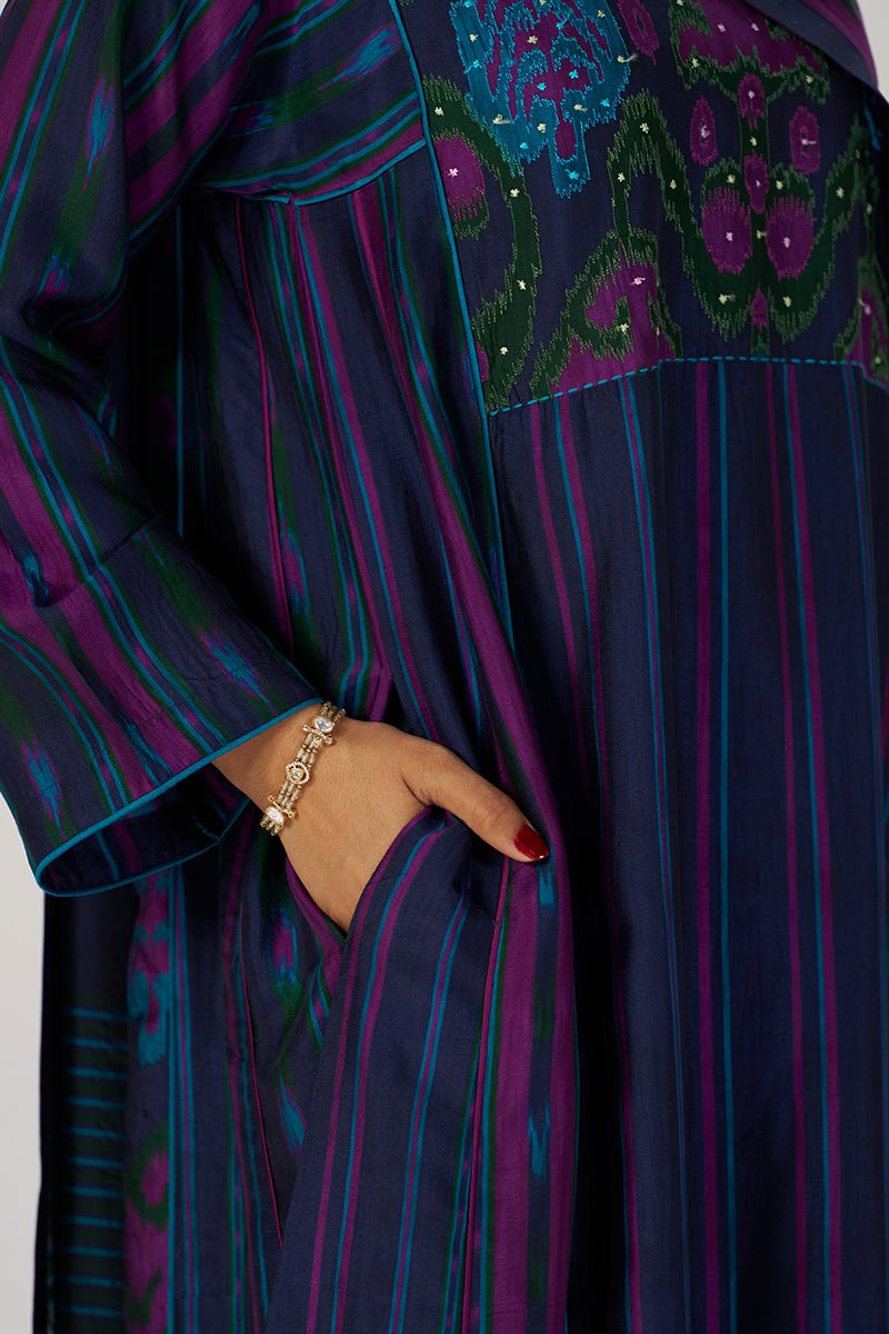 A close-up of a long-sleeved tunic or abaya with vertical stripes in deep purple, navy, and teal, featuring a decorative embroidered yoke with floral motifs and a hand tucked into the side pocket. | Medieval Blue Silk Festive Kurta Set - Ganga Fashions