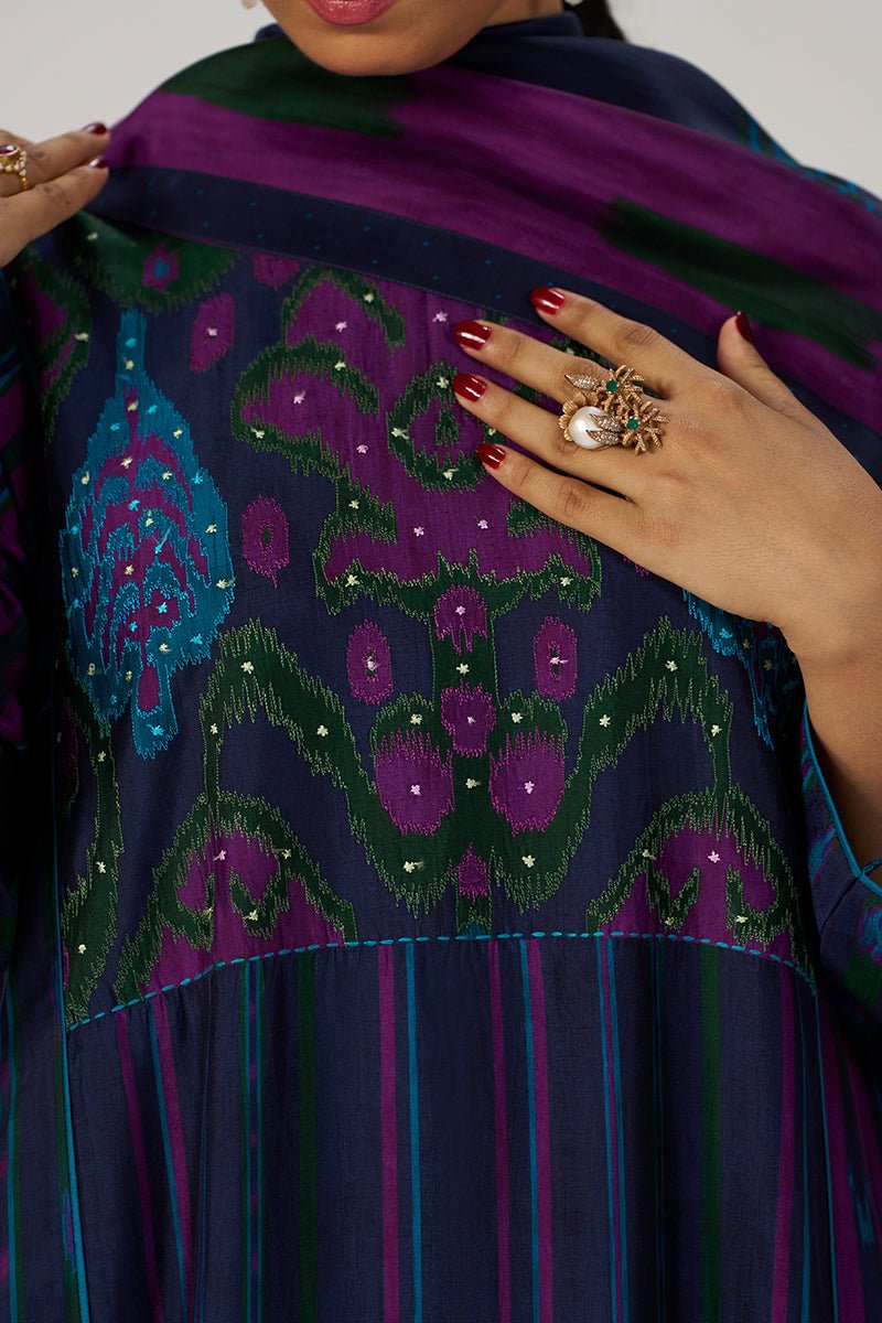 Close-up of a woman wearing a dark navy embroidered dress with purple and green patterns, holding a matching purple dupatta. She is wearing a large statement ring with a pearl and green stones. | Medieval Blue Silk Festive Kurta Set - Ganga Fashions
