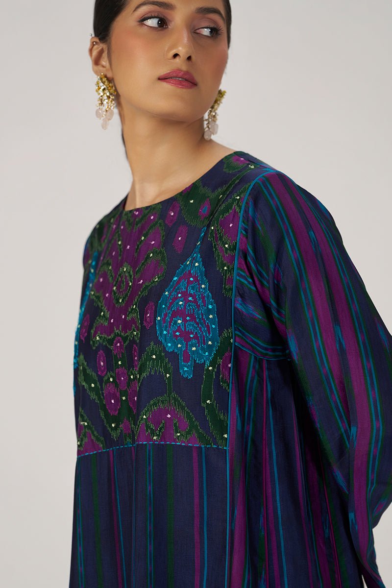 A woman wearing a dark purple and navy blue embroidered tunic top with green paisley patterns and blue vertical stripes on the sleeves, shown in a close-up profile against a light gray background. | Medieval Blue Silk Festive Kurta Set - Ganga Fashions