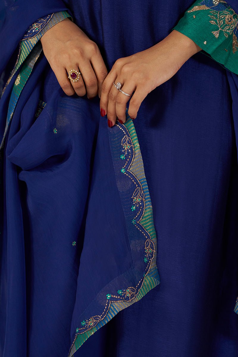 Close-up of a person wearing a royal blue fabric garment with a sheer green and blue embroidered border, holding the fabric at the waist. The person's hands are visible, one with a red gemstone ring and the other with a silver ring. | Space Blue Festive Kurta Set - Ganga Fashions