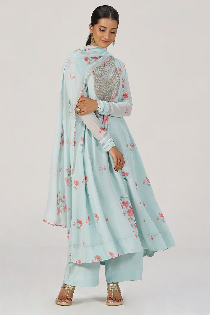 A woman modeling a light blue traditional outfit with pink floral prints, featuring a long kurta, matching pants, and a sheer dupatta with a silver embroidered yoke, set against a plain light gray background. | Whispering Blue Georgette Festive Kurta Set - Ganga Fashions