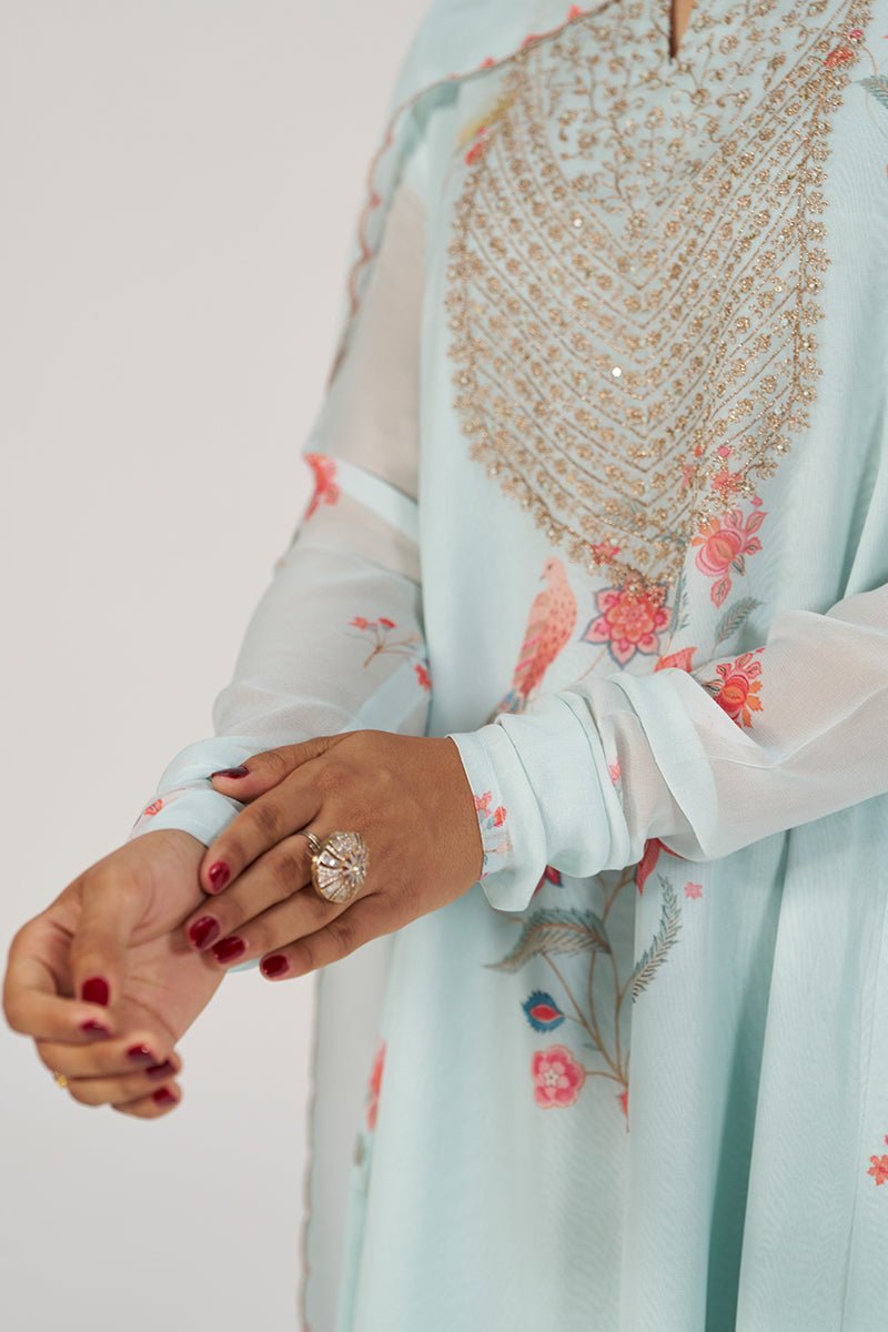 A close-up of a person wearing a light blue embroidered traditional outfit with sheer long sleeves, floral prints, and a large ornate gold embellished neckline. The person is shown from the chest down against a plain light background, with one hand resting on the other and wearing a ring. | Whispering Blue Georgette Festive Kurta Set - Ganga Fashions