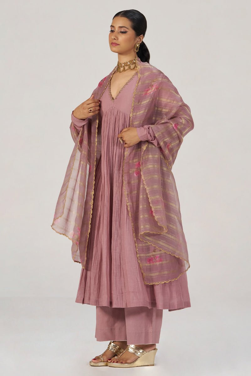 A woman models a dusty pink traditional outfit consisting of a pleated kurta, matching trousers, and a sheer striped dupatta with gold borders, set against a plain light gray background. | Woodrose Chanderi Silk Festive Kurta Set - Ganga Fashions