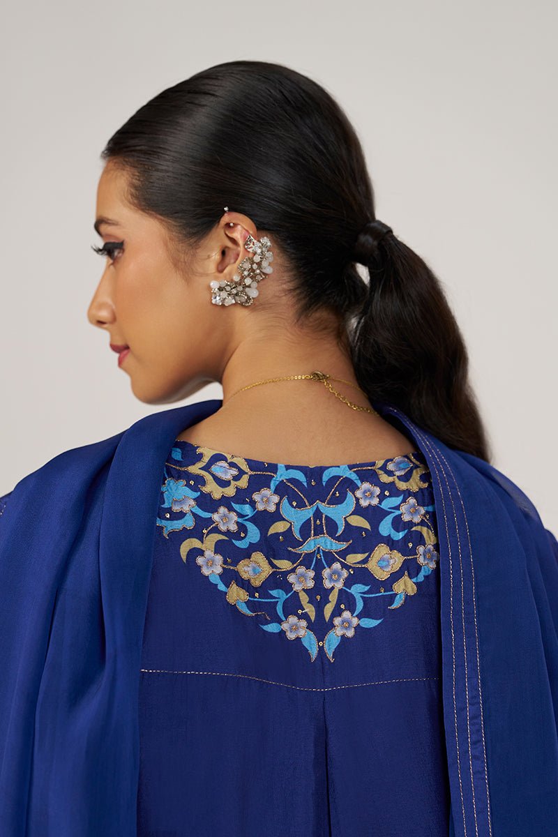 A woman shown from the back wearing a royal blue traditional outfit with a heavily embroidered floral yoke in blue, gold, and white. She is wearing a matching blue shawl and ornate silver earrings against a plain light gray studio background. | Royal Blue Festive Kurta Set - Ganga Fashions