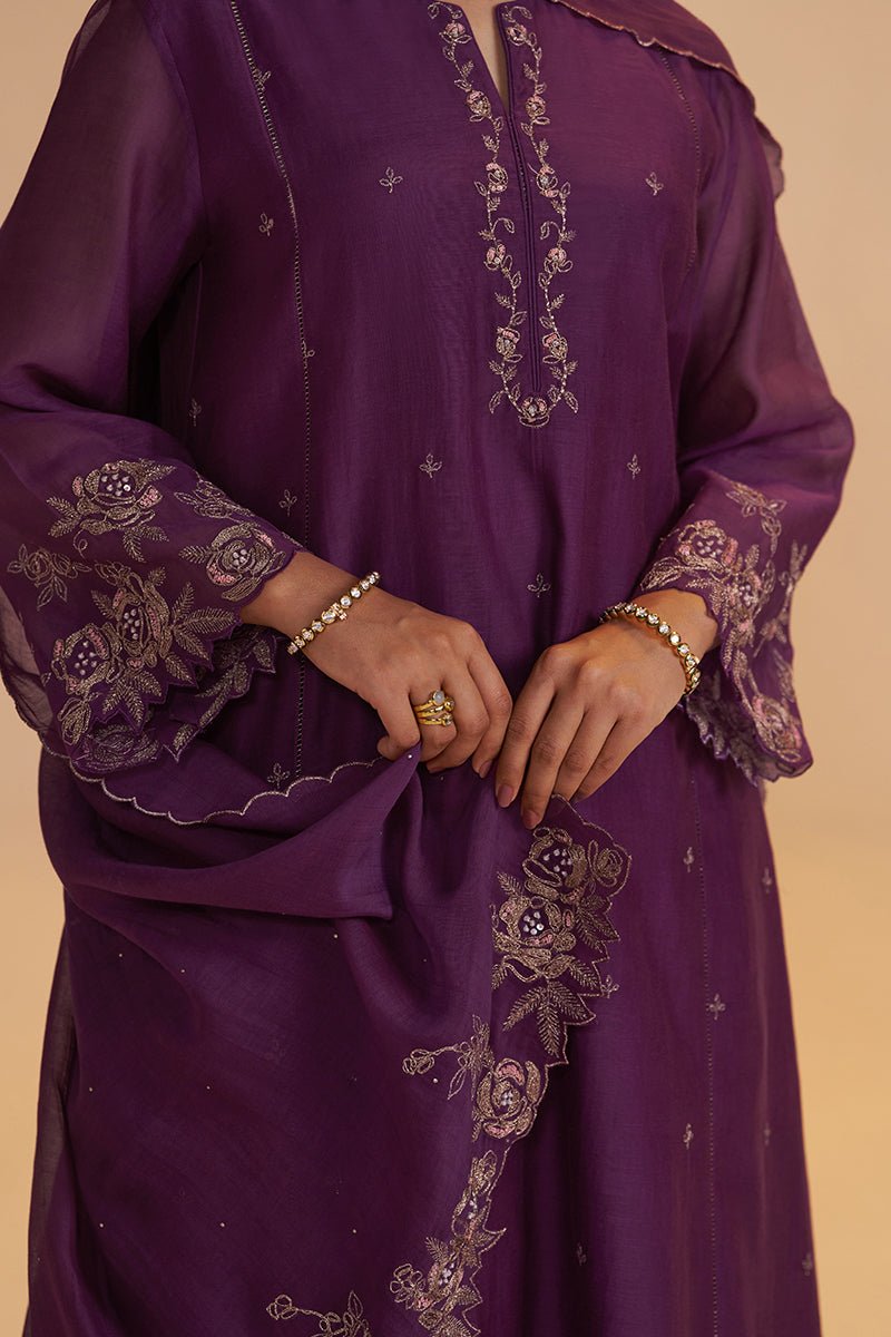A close-up of a person wearing a purple traditional embroidered tunic with floral lace detailing on the sleeves and hem, paired with gold jewelry against a warm beige background. | Purple Festive Kurta Set - Ganga Fashions
