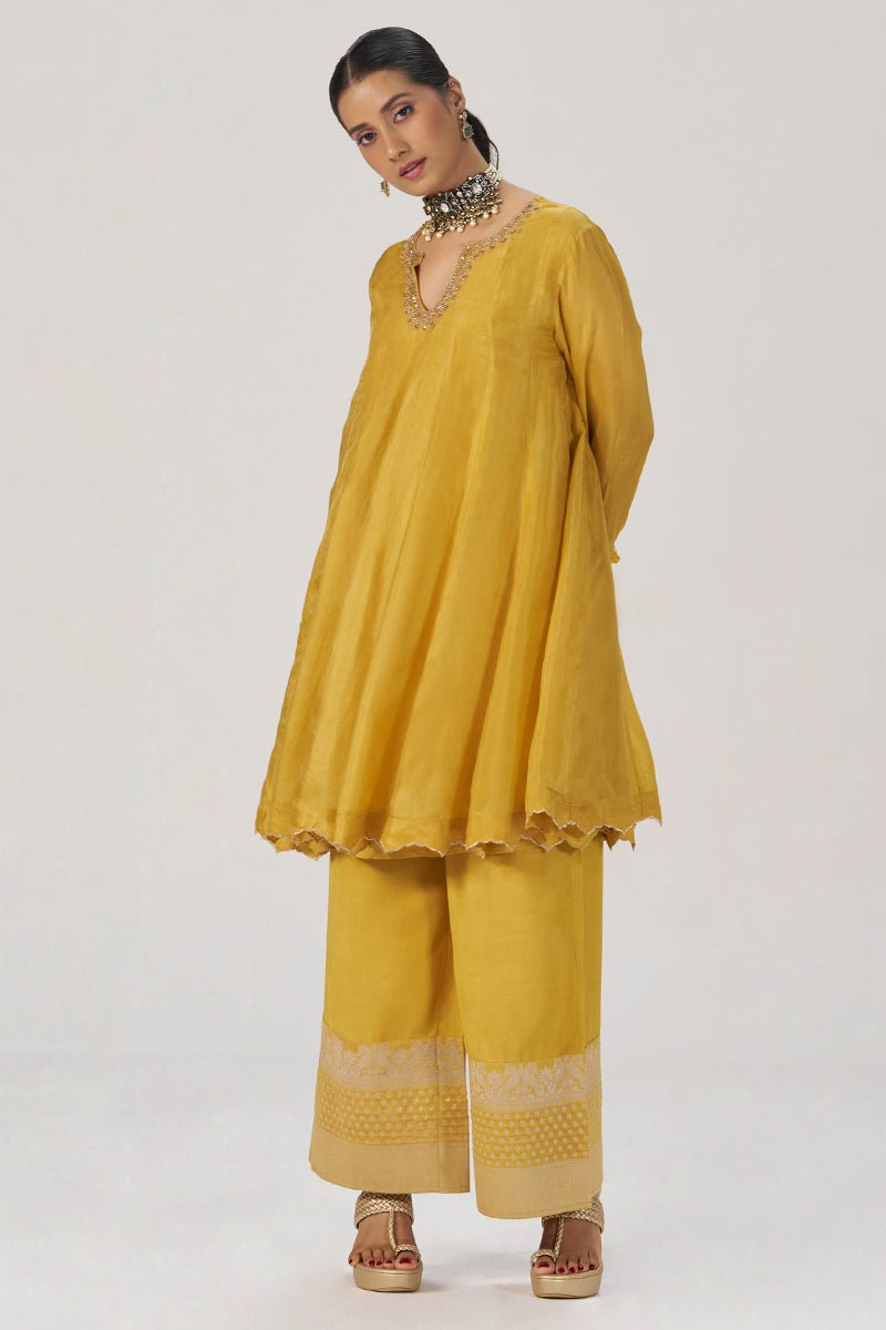 A woman modeling a mustard yellow traditional three-piece outfit consisting of a long-sleeve kurta with scalloped hem, matching wide-leg trousers with embroidered hems, and a statement necklace, set against a plain light gray background. | Golden Yellow Festive Silk Co-ord Set - Ganga Fashions