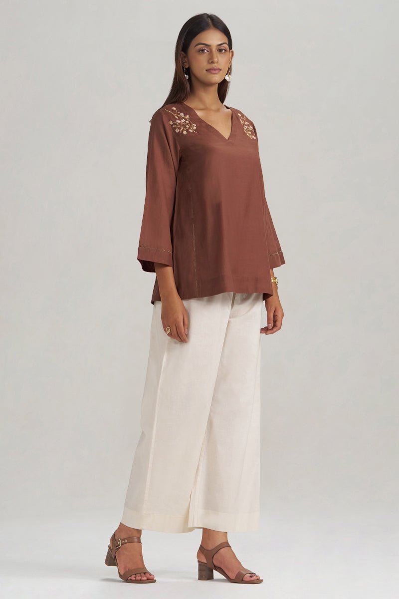 A woman modeling a rust-brown V-neck top with floral embroidery on the shoulders, paired with off-white wide-leg trousers and brown heeled sandals, against a light gray studio background. | Cocoa Brown Embroidered Mul-Mul Tunic - Ganga Fashions