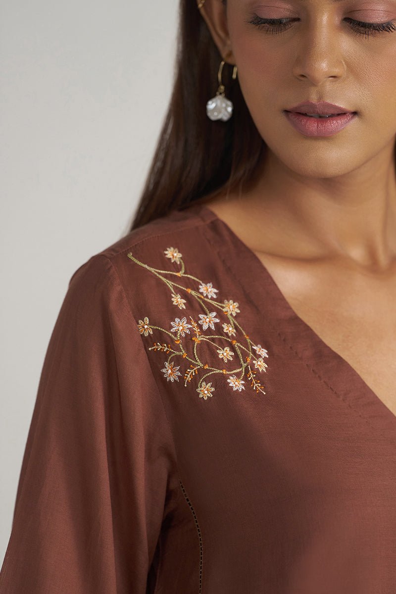 Close-up of a woman wearing a brown V-neck top with delicate floral embroidery on the shoulder, paired with dangling earrings, against a plain light background. | Cocoa Brown Embroidered Mul-Mul Tunic - Ganga Fashions