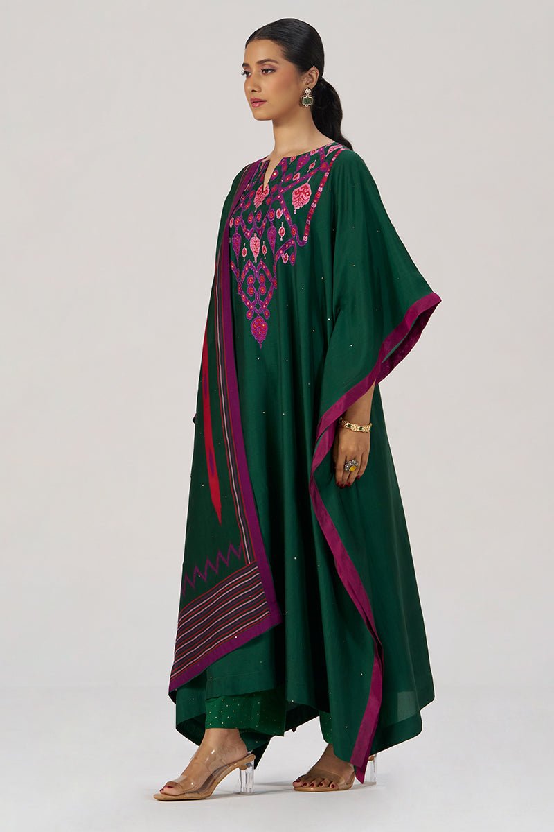 A woman modeling a dark green traditional outfit with magenta and pink embroidered details on the chest, wide draped sleeves, and a matching dupatta with geometric borders, photographed against a light gray studio background. | Evergreen Silk Festive Kaftaan Set - Ganga Fashions
