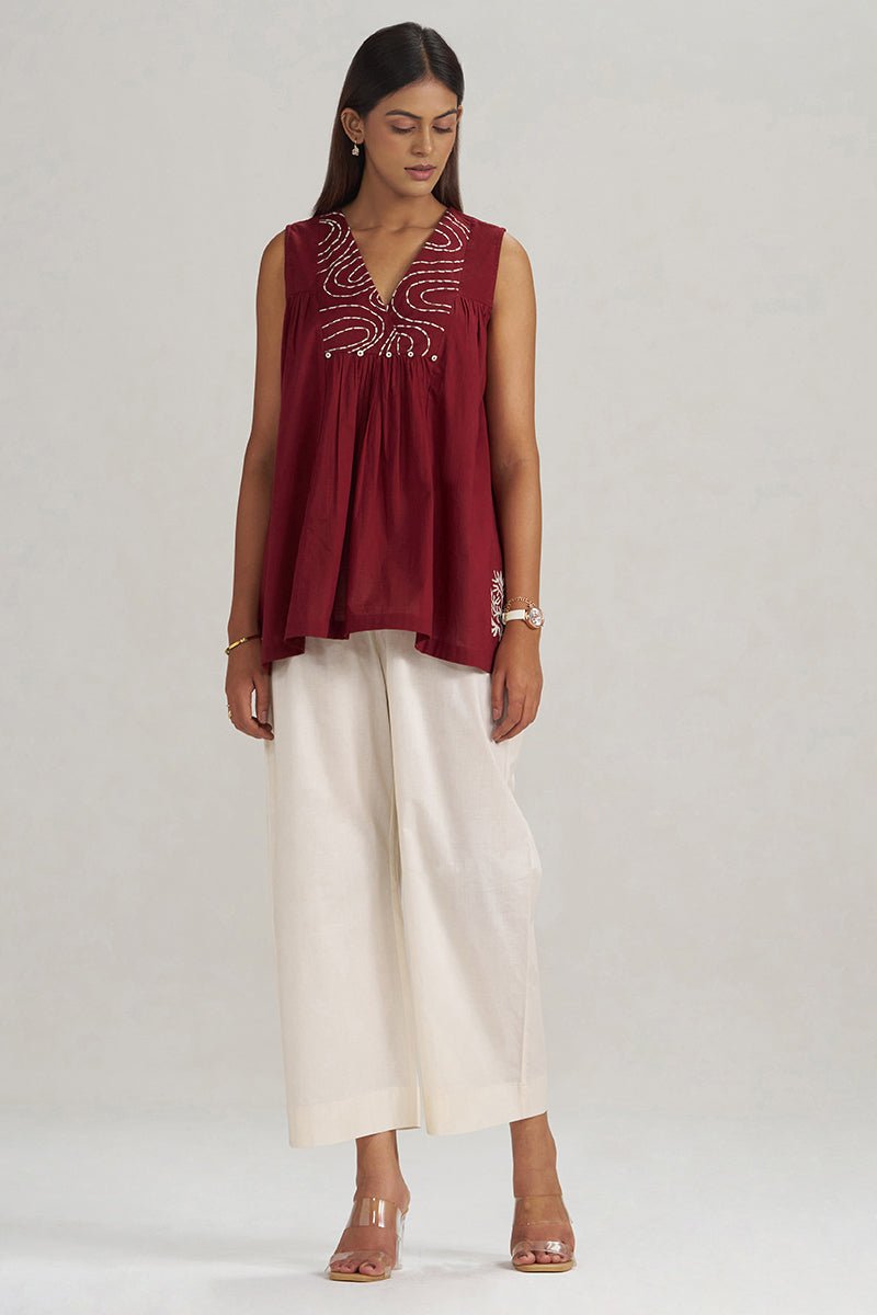 A woman modeling a sleeveless deep maroon top with silver beaded embroidery around the V-neckline, paired with off-white wide-leg cropped pants and clear high-heeled sandals, against a light gray studio background. | Maroon Embroidered Mul-Mul Tunic - Ganga Fashions