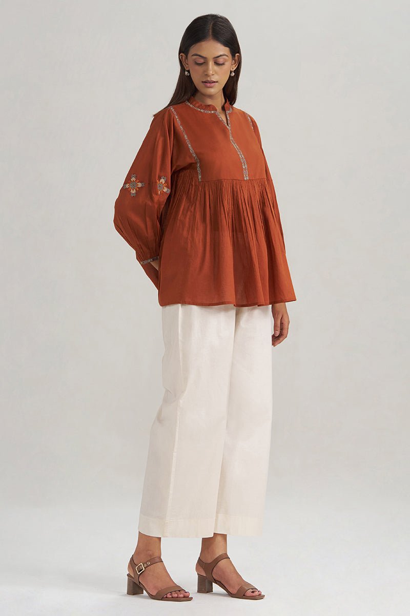 A woman modeling a rust-orange long-sleeve peplum top with embroidered details on the chest and sleeves, paired with off-white cropped trousers and brown heeled sandals, against a light gray studio background. | Rust Embroidered Mul-Mul Tunic - Ganga Fashions