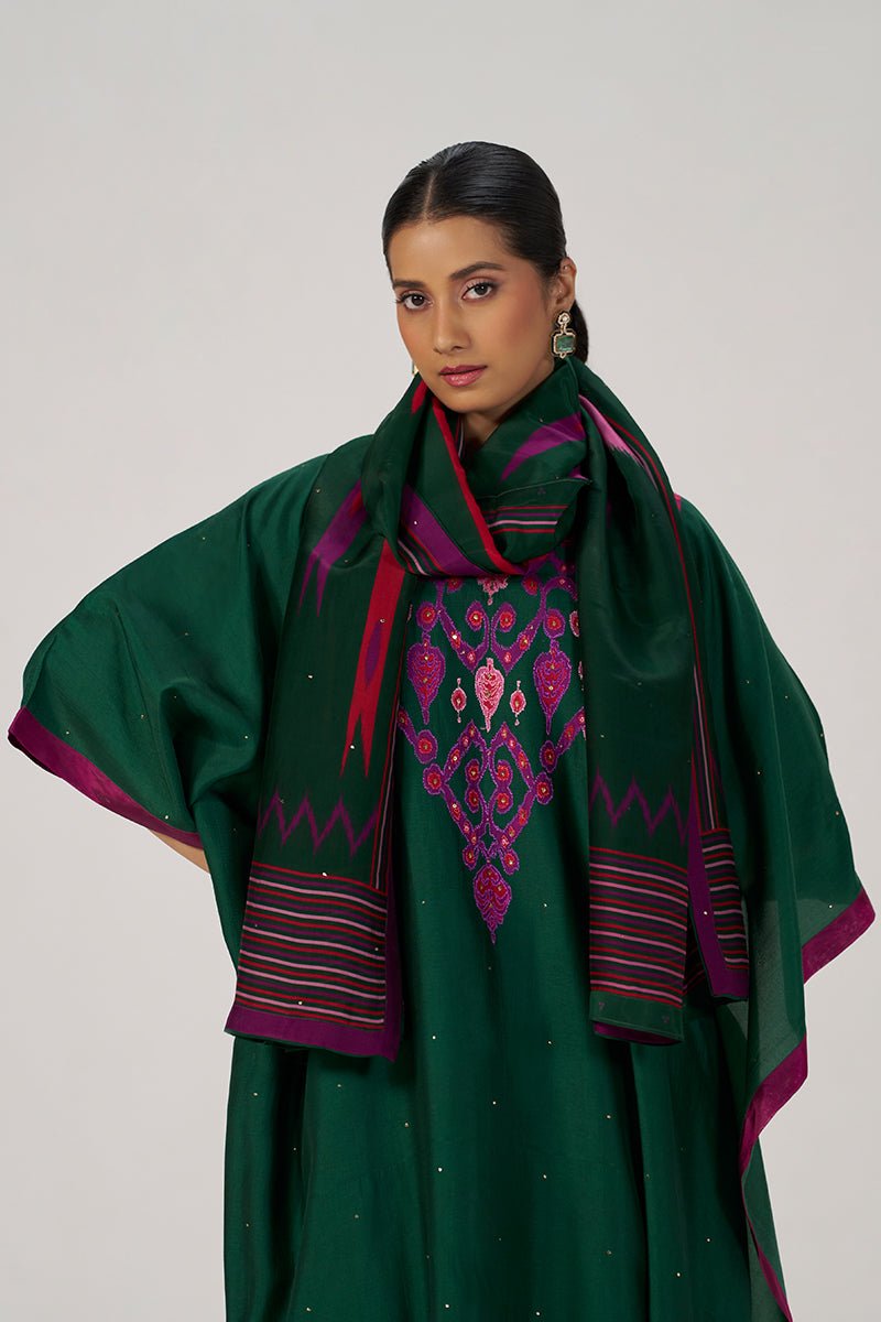 A woman modeling a dark green traditional outfit with a matching scarf draped over her shoulders. The outfit features magenta and purple embroidered details on the chest and striped patterned borders on the sleeves and scarf. | Evergreen Silk Festive Kaftaan Set - Ganga Fashions