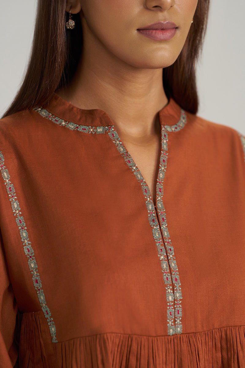 Close-up of a woman wearing a rust-orange tunic top with detailed multicolor embroidery along the split neckline and a fringed hem. | Rust Embroidered Mul-Mul Tunic - Ganga Fashions