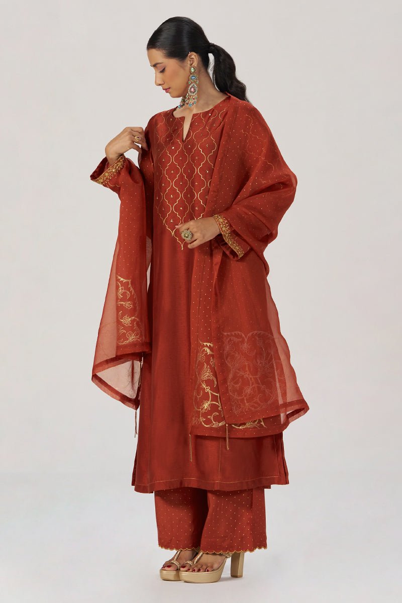 A woman modeling a rust-red traditional South Asian outfit with gold embroidery, matching pants, and a sheer dupatta, photographed against a plain light gray studio background. | Rust Raw Silk Festive Kurta Set - Ganga Fashions