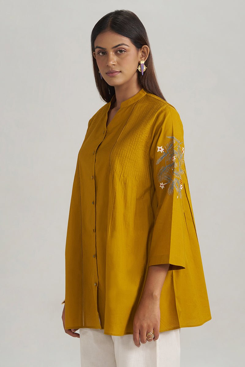 A woman modeling a mustard yellow button-up kurta with white pants, standing against a light gray studio background. The kurta features a textured fabric, three-quarter sleeves, and delicate floral embroidery on the upper sleeve. | Mustard Mul-Mul Embroidered Tunic - Ganga Fashions