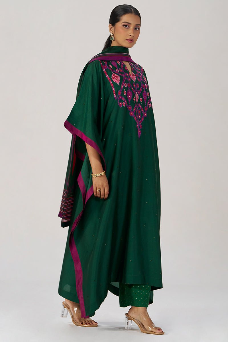 A woman models a dark green traditional outfit with magenta and pink embroidered details on the chest, paired with matching green pants and a sheer green dupatta with magenta borders, against a light gray studio background. | Evergreen Silk Festive Kaftaan Set - Ganga Fashions