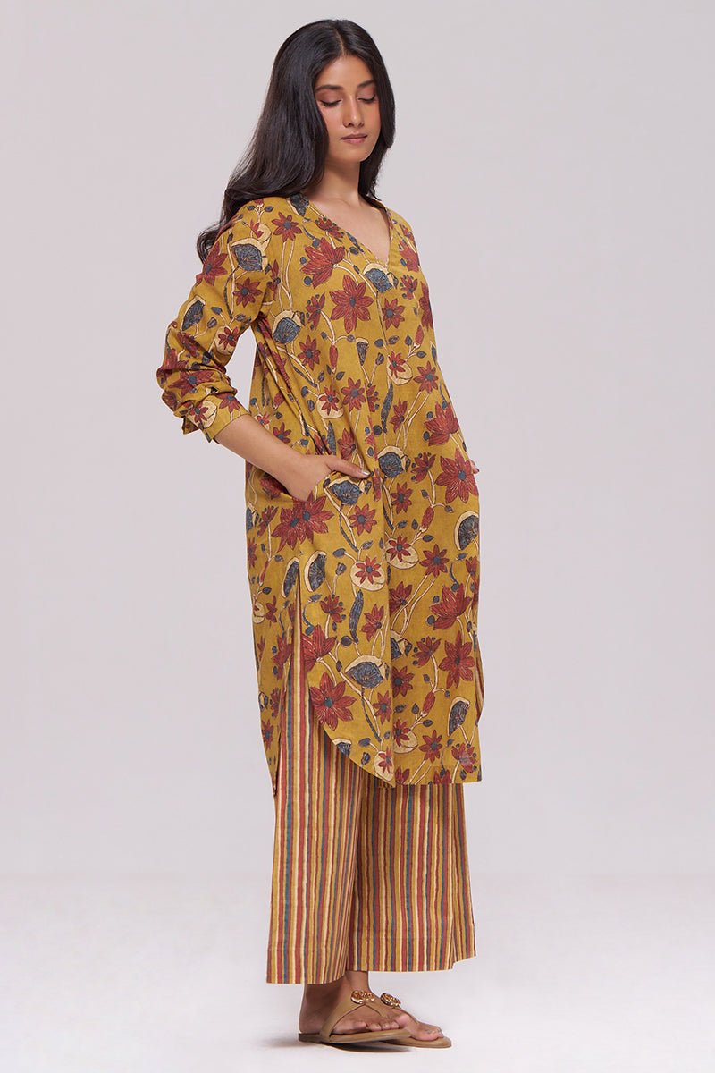 A woman modeling a mustard yellow kurta with a red, green, and blue floral print, paired with matching striped pants, against a light gray studio background. | Mustard and Brick Red Floral Kurta and Stripe Bottom Set - Ganga Fashions