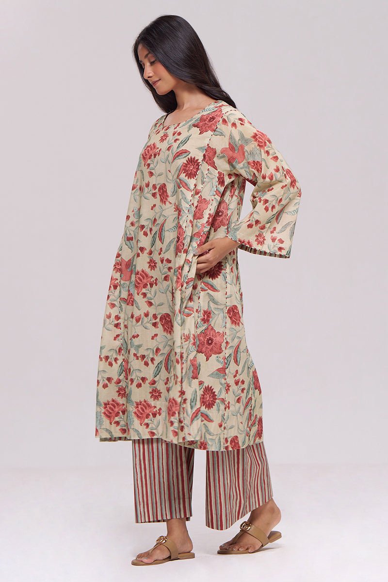 A woman modeling a long cream-colored kurta with a red and green floral print, paired with red and beige striped pants, against a plain light gray studio background. | Vintage Beige and Rose Floral Print Kurta and Stripe Bottom Set - Ganga Fashions