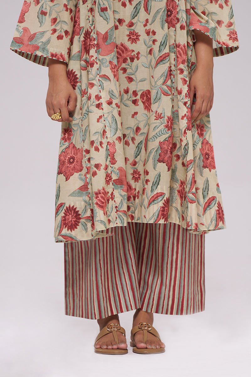 A model wearing a cream-colored tunic with a red and green floral print, paired with red and green striped pants and brown sandals, shown from the waist down against a plain white background. | Vintage Beige and Rose Floral Print Kurta and Stripe Bottom Set - Ganga Fashions