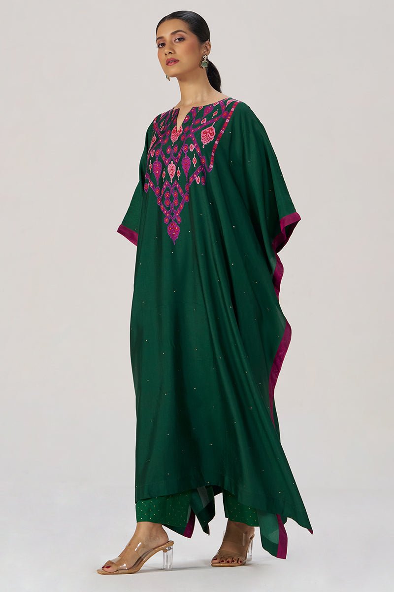 A woman models a dark green kaftan-style dress with magenta and pink embroidered detailing around the neckline and a matching magenta border along the sleeves and hem, photographed against a plain light gray studio background. | Evergreen Silk Festive Kaftaan Set - Ganga Fashions