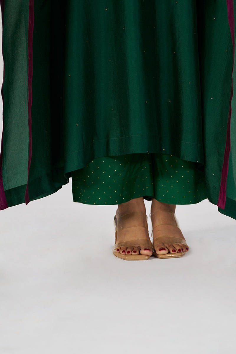 A close-up of a person wearing a dark green traditional outfit with gold dot patterns and a sheer green dupatta, paired with tan leather slide sandals, shown from the waist down on a white studio background. | Evergreen Silk Festive Kaftaan Set - Ganga Fashions