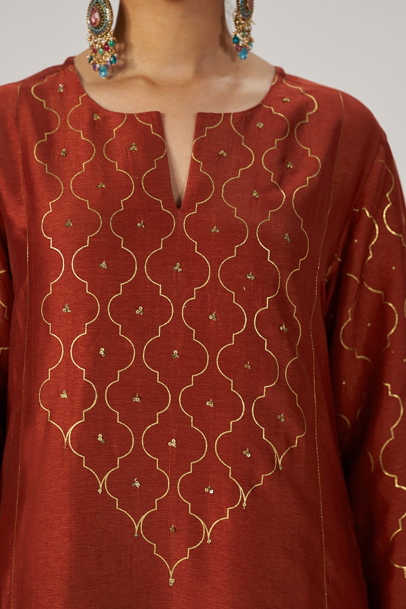 A close-up of a rust-red or terracotta-colored traditional tunic with gold geometric embroidery and small metallic embellishments, shown on a model wearing colorful earrings. | Rust Raw Silk Festive Kurta Set - Ganga Fashions
