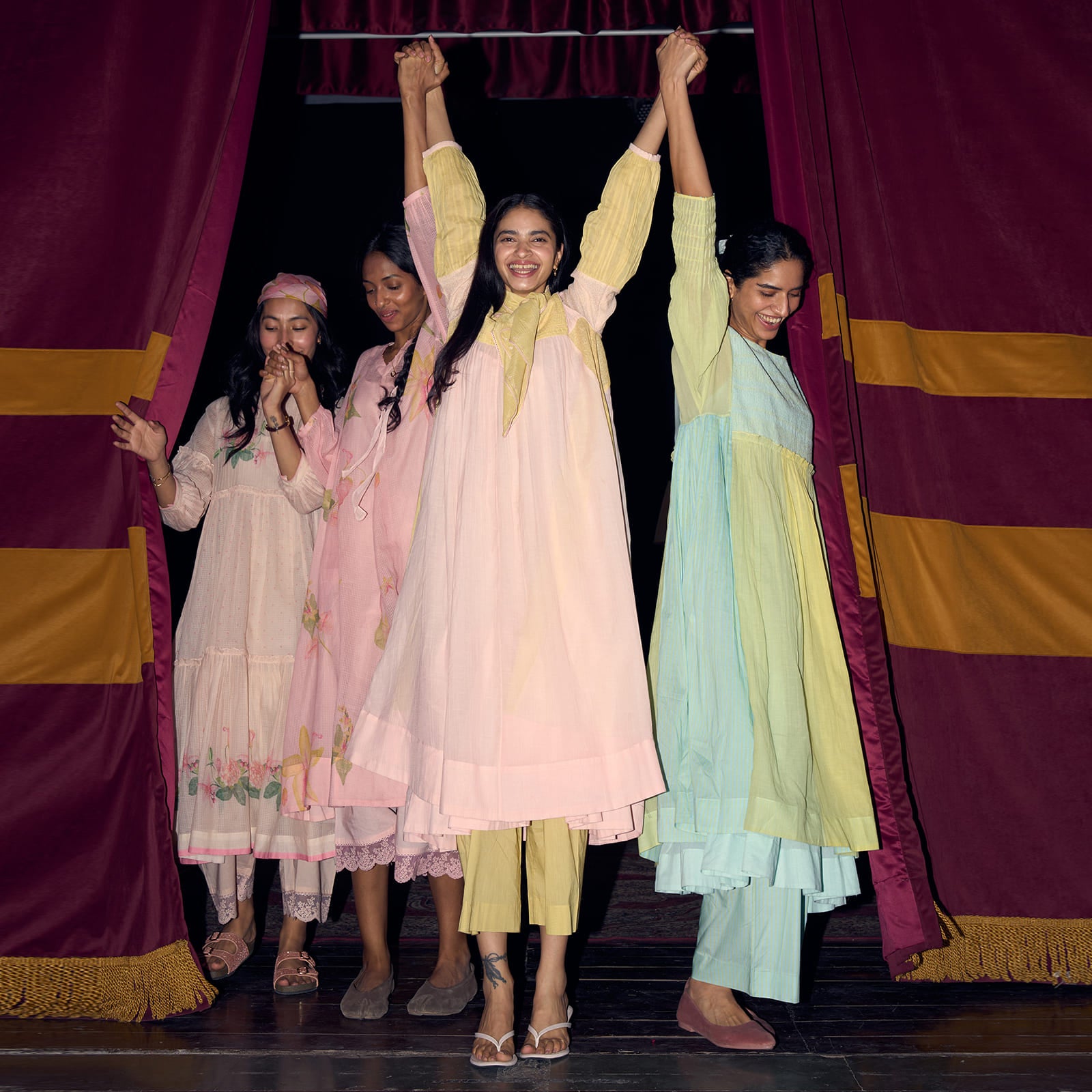 Four women in colorful traditional outfits standing on a stage with a red curtain backdrop. | rangmanch collection | elegant kurta sets & co-ords | ganga fashion