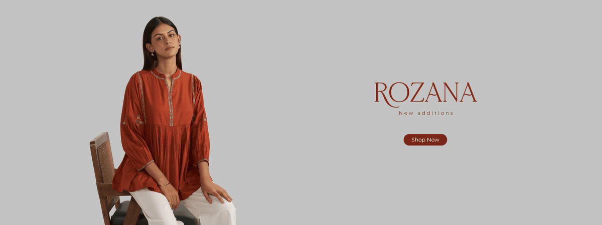 A woman seated on a wooden chair wearing a rust-orange embroidered kurta and white trousers, set against a light gray background with the brand name ROZANA and a 'Shop Now' button on the right. | ROZANA Collection Ganga