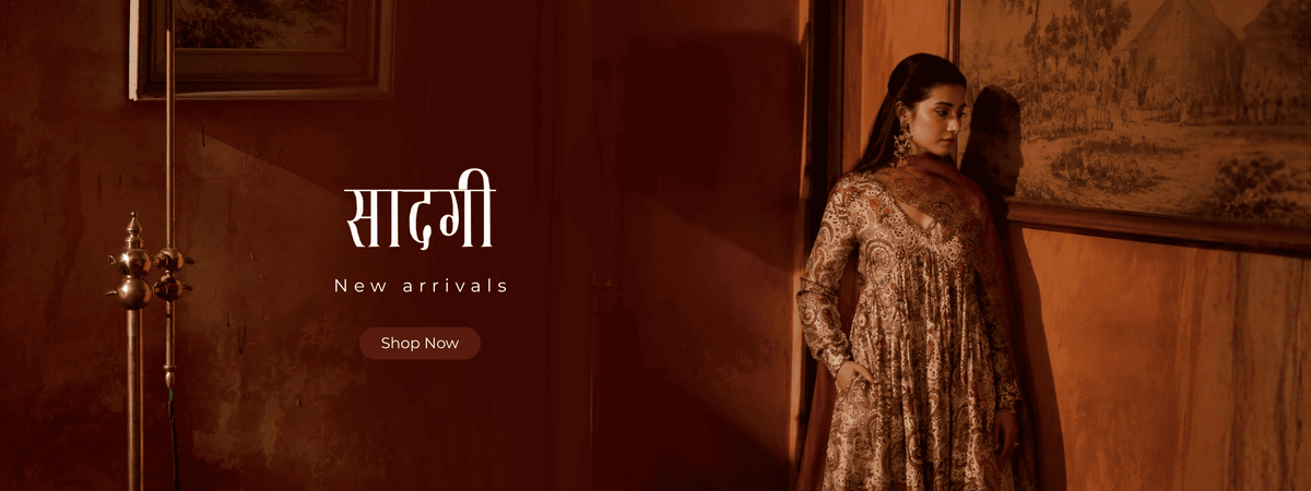 A woman wearing a traditional Indian salwar kameez outfit with a patterned beige and brown tunic and a deep purple dupatta, standing in a dimly lit room with warm brown walls and framed art. | Ganga Saadagi Collections