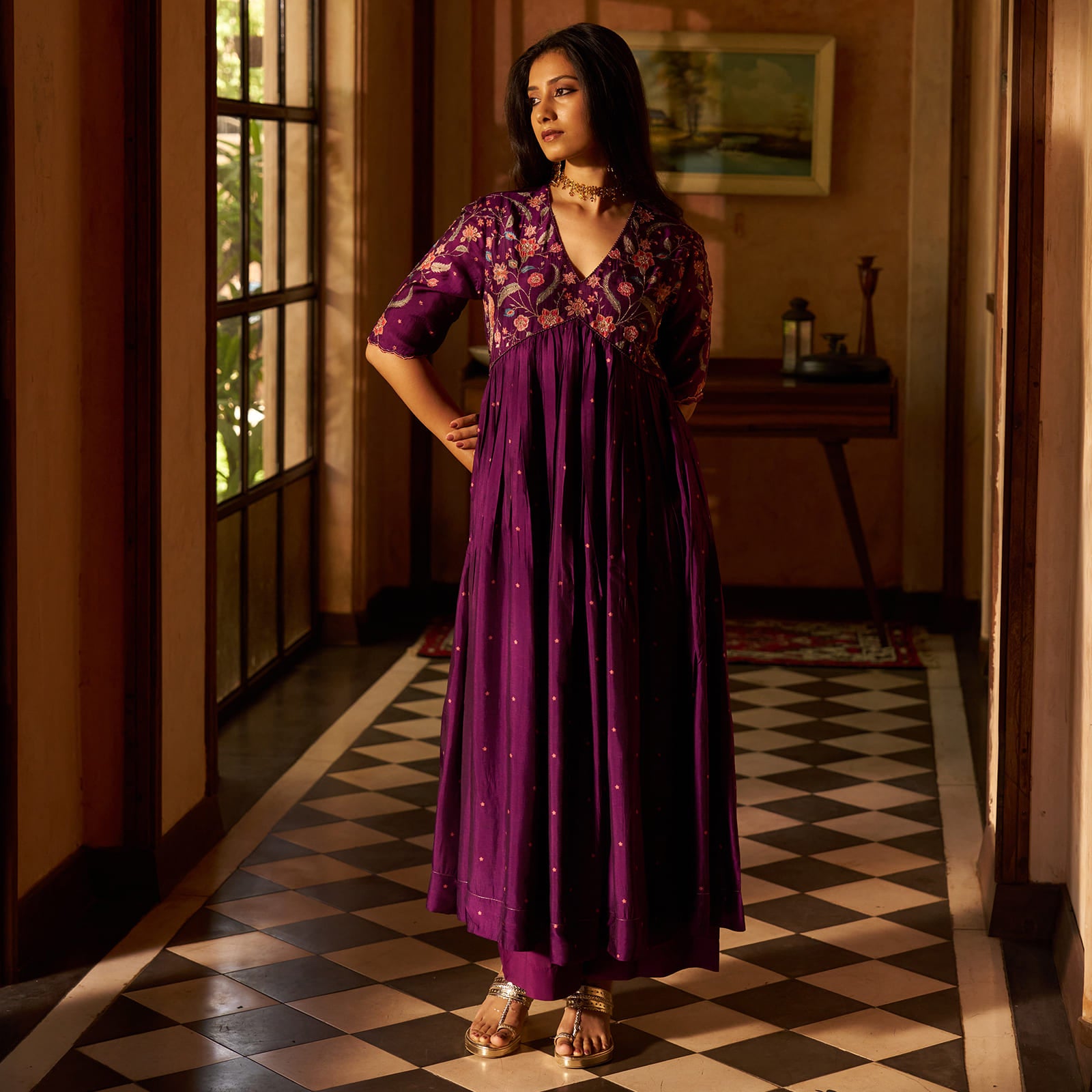 A woman models a purple traditional Indian dress with floral embroidery on the bodice and scattered gold zari embellishments on the skirt, standing in a hallway with a checkered floor. |