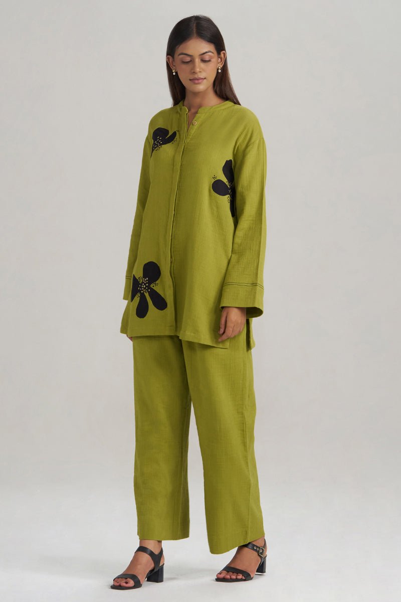 A woman modeling a matching olive green two-piece set consisting of a long-sleeve button-front tunic with black floral applique and wide-leg trousers, paired with black heeled sandals, against a light gray studio background. | Lime Green Botanical Co-ord Set - Ganga Fashions