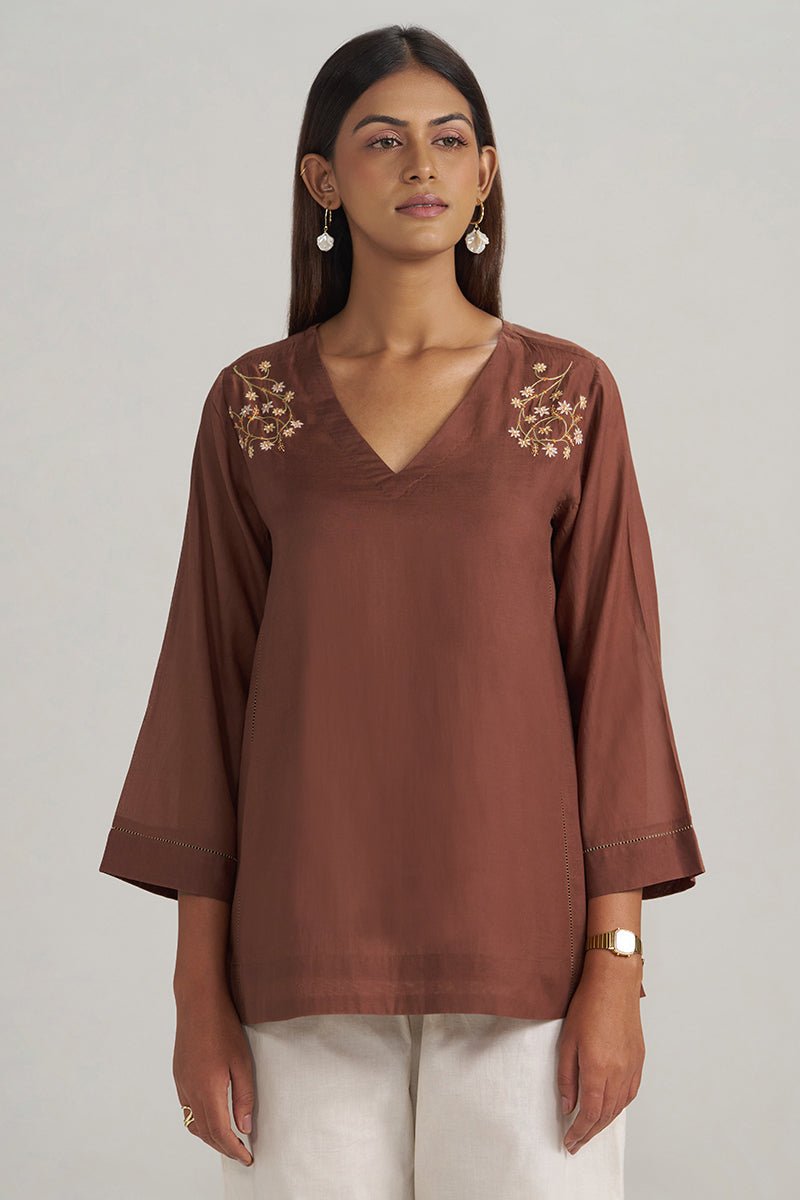 A woman modeling a brown V-neck top with gold floral embroidery on the shoulders, paired with white pants, against a plain light gray background. | Cocoa Brown Embroidered Mul-Mul Tunic - Ganga Fashions