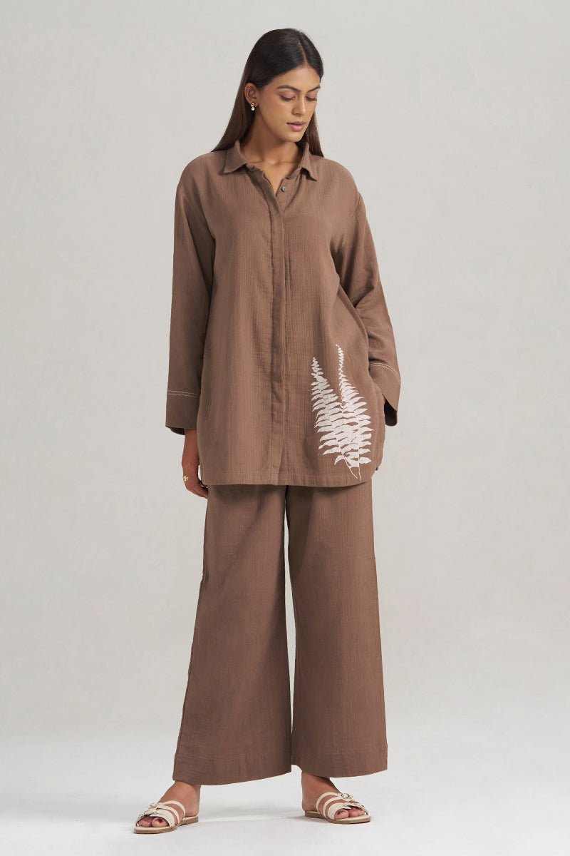 A woman modeling a brown two-piece linen set consisting of a long-sleeve button-up shirt and matching wide-leg trousers, featuring a white fern leaf embroidery on the shirt pocket. | Chocolate and Fern Co-ord Set - Ganga Fashions