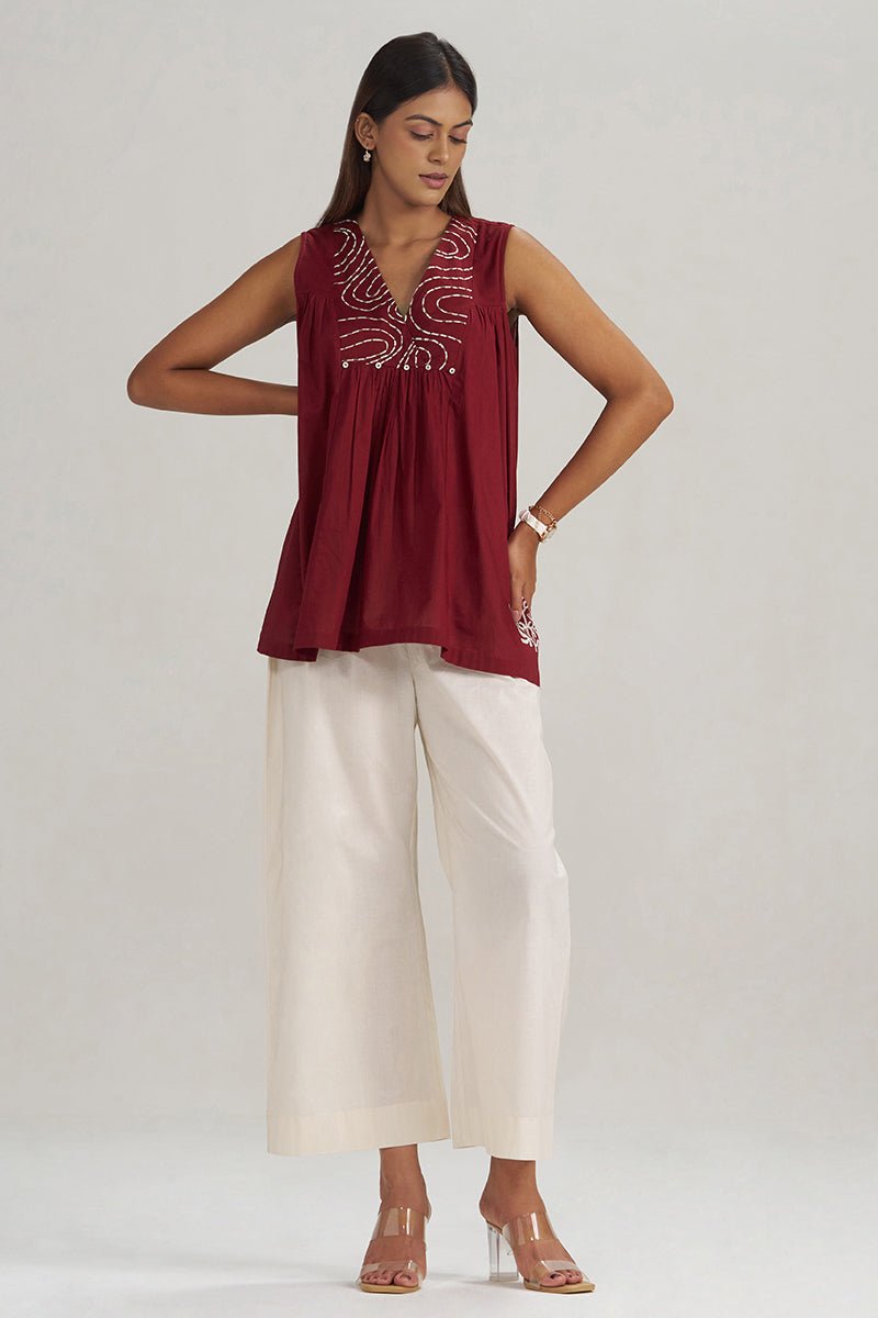A woman modeling a sleeveless maroon top with silver embroidery on the chest, paired with white cropped pants and clear high-heeled sandals, against a light gray studio background. | Maroon Embroidered Mul-Mul Tunic - Ganga Fashions