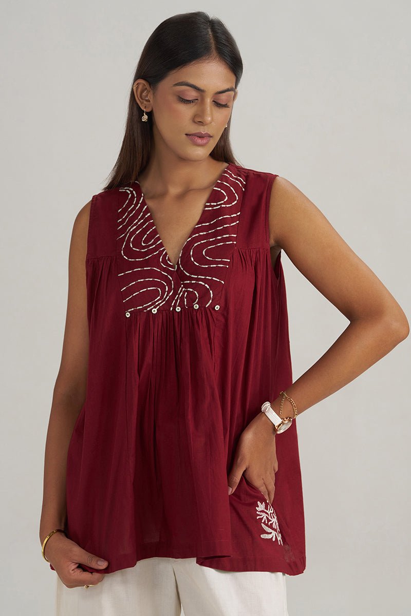 A woman wearing a sleeveless deep maroon top with white embroidered patterns on the chest and a matching embroidered detail on the lower left side, paired with white pants, against a light gray background. | Maroon Embroidered Mul-Mul Tunic - Ganga Fashions