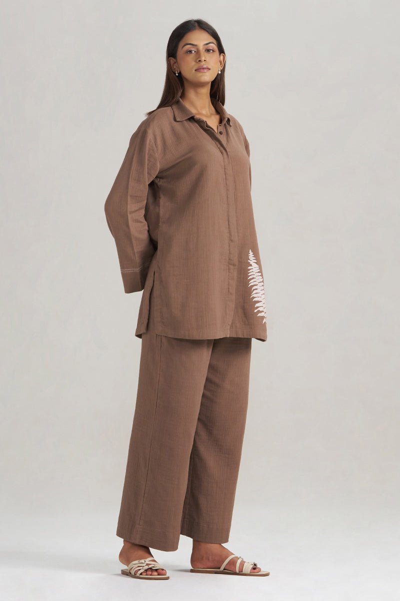 A woman modeling a matching brown two-piece linen set consisting of a long-sleeve button-up shirt with white embroidered fern motifs on the sleeve and pants, paired with beige strappy sandals, against a light gray studio background. | Chocolate and Fern Co-ord Set - Ganga Fashions