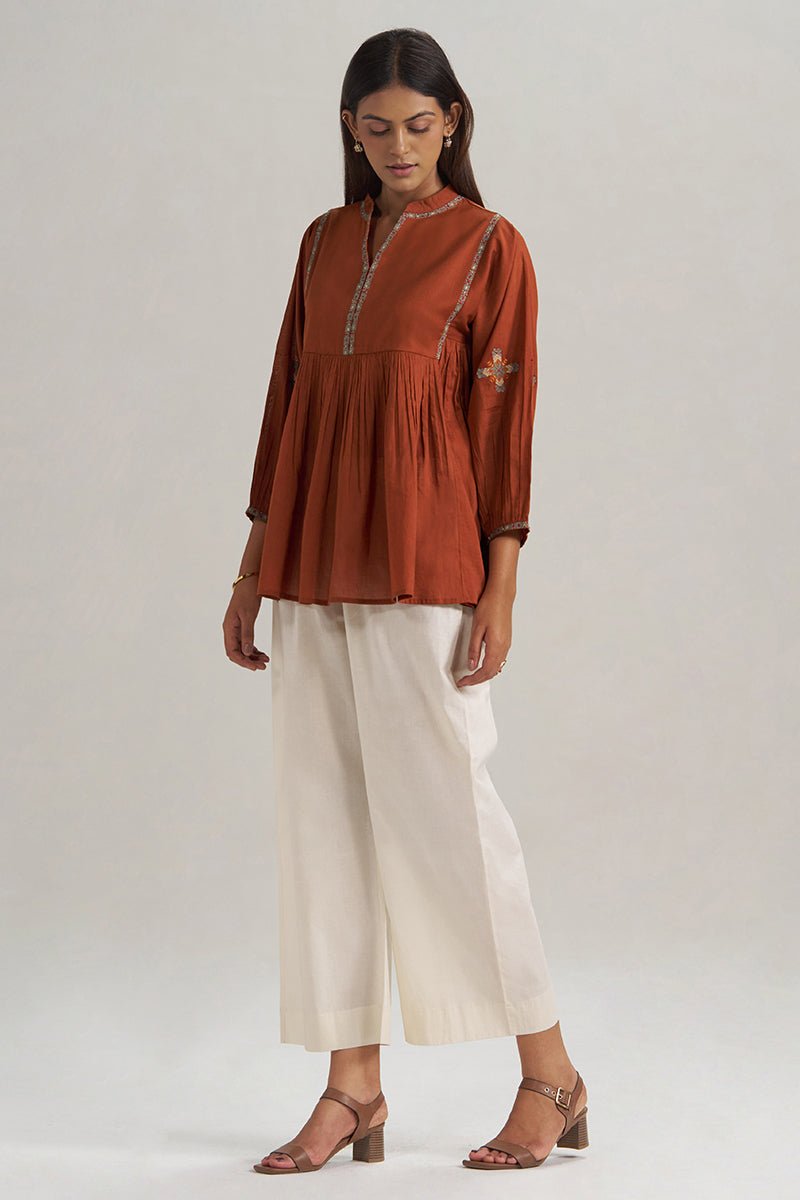 A woman modeling a rust-orange peplum kurta with embroidered details, paired with off-white cropped trousers and brown heeled sandals, against a neutral light gray background. | Rust Embroidered Mul-Mul Tunic - Ganga Fashions