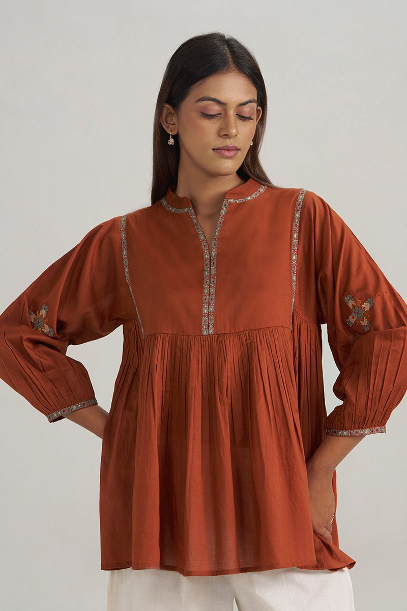 A woman wearing a rust-orange cotton kurta with silver embroidered trim and sleeve details, paired with white pants, photographed against a neutral light gray background. | Rust Embroidered Mul-Mul Tunic - Ganga Fashions