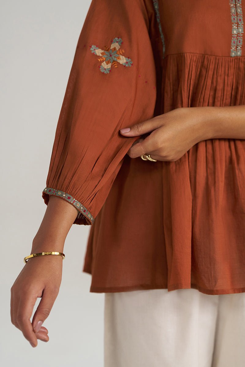 Close-up of a terracotta-colored cotton kurta with embroidered details on the sleeve and a pleated bodice, worn by a model with white trousers. | Rust Embroidered Mul-Mul Tunic - Ganga Fashions