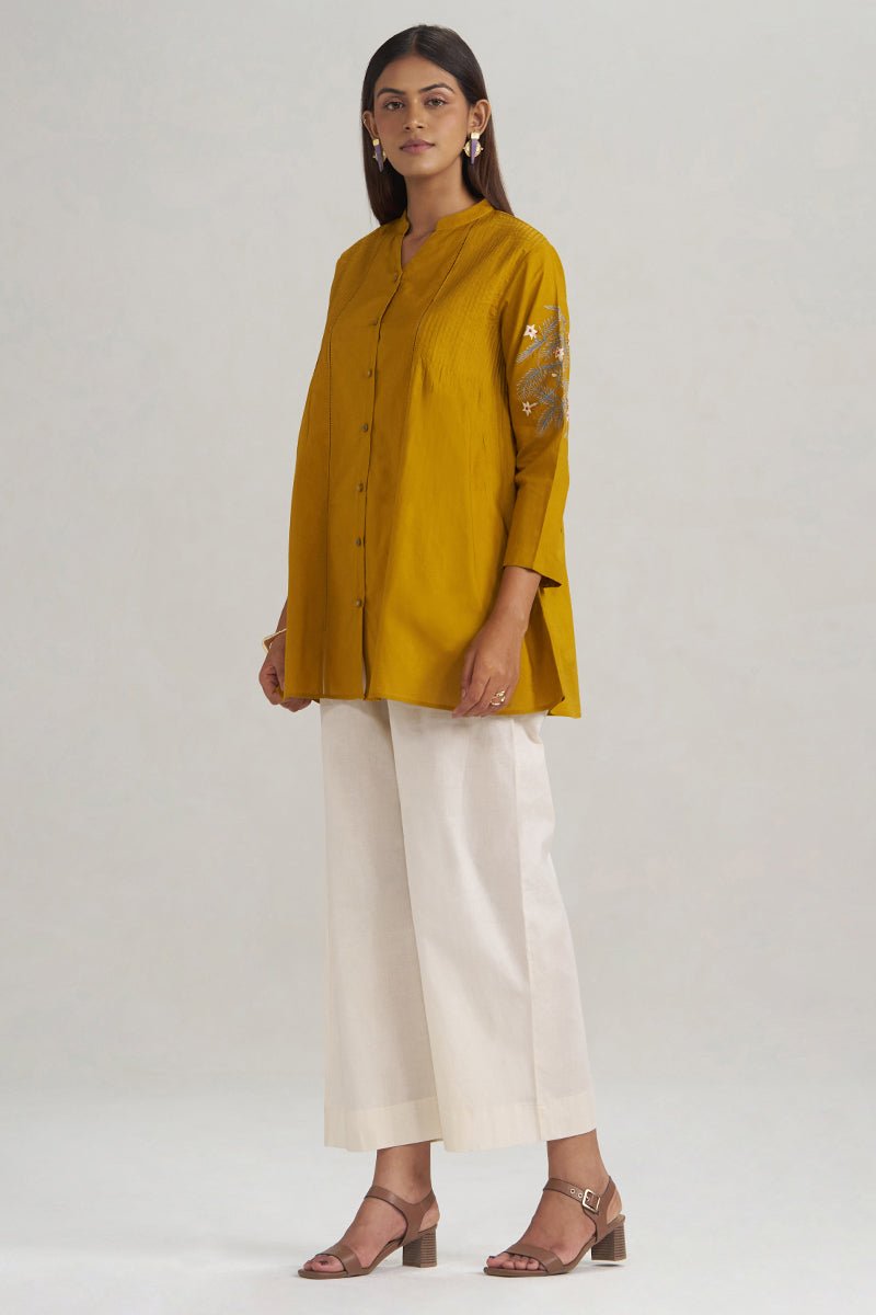 A woman modeling a mustard yellow button-up kurta with gold embroidery on the sleeve, paired with off-white cropped trousers and brown heeled sandals, against a light gray studio background. | Mustard Mul-Mul Embroidered Tunic - Ganga Fashions