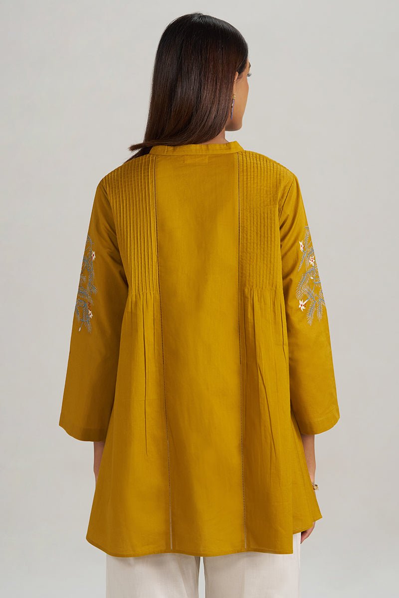 A woman viewed from the back wearing a mustard yellow tunic-style top with vertical pleats and embroidered details on the sleeves, paired with white pants against a light gray studio background. | Mustard Mul-Mul Embroidered Tunic - Ganga Fashions