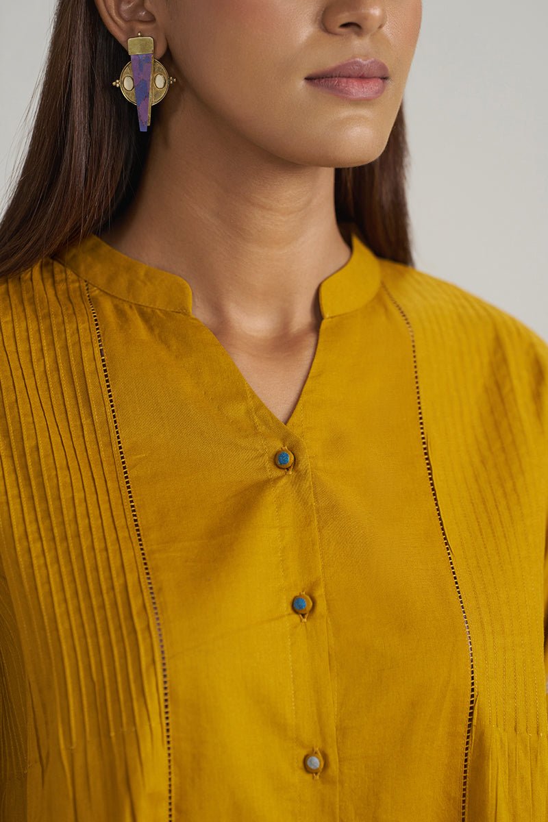 Close-up of a woman wearing a mustard yellow pleated kurta with a mandarin collar and decorative buttons, paired with a gold and purple earring. | Mustard Mul-Mul Embroidered Tunic - Ganga Fashions