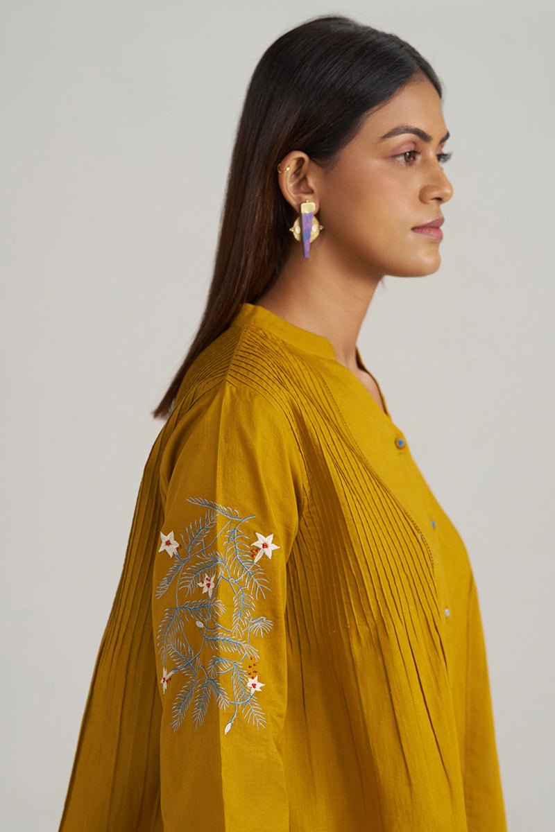 A woman shown in profile wearing a mustard yellow embroidered kurta with pleated detailing on the front and sleeve. The kurta features white and light blue floral embroidery on the upper sleeve. | Mustard Mul-Mul Embroidered Tunic - Ganga Fashions