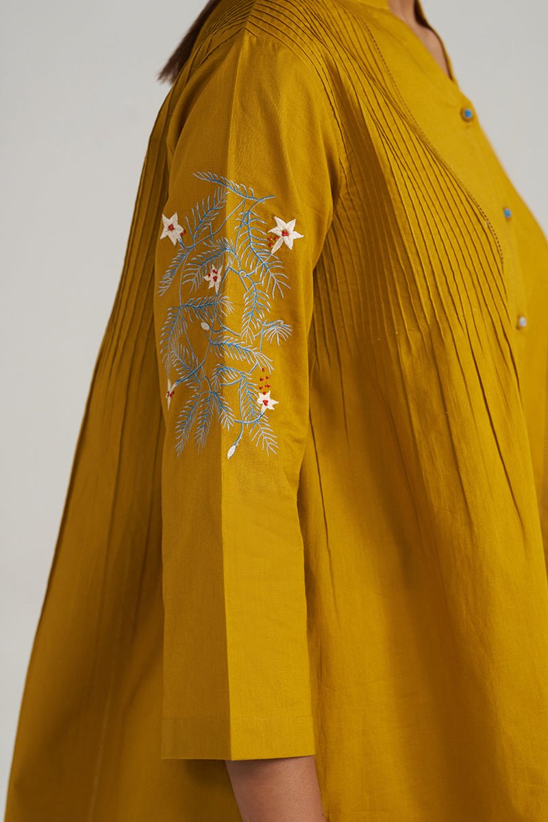 Close-up of a mustard yellow pleated kurta or tunic with embroidered floral and leaf motifs on the sleeve, shown on a model against a light gray background. | Mustard Mul-Mul Embroidered Tunic - Ganga Fashions